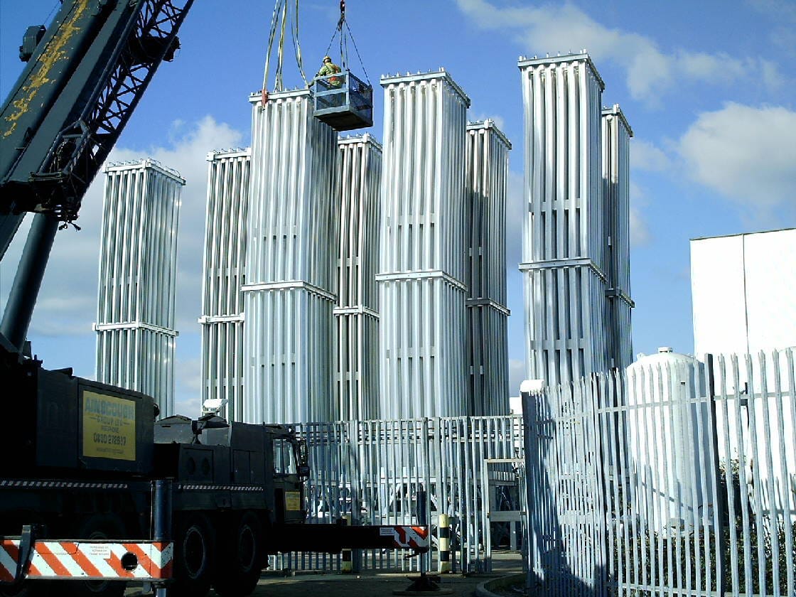 A field of tall finned-aluminum FWM ambient air argon vaporizer banks being installed on site with a mobile crane lifting a technician alongside the stacks