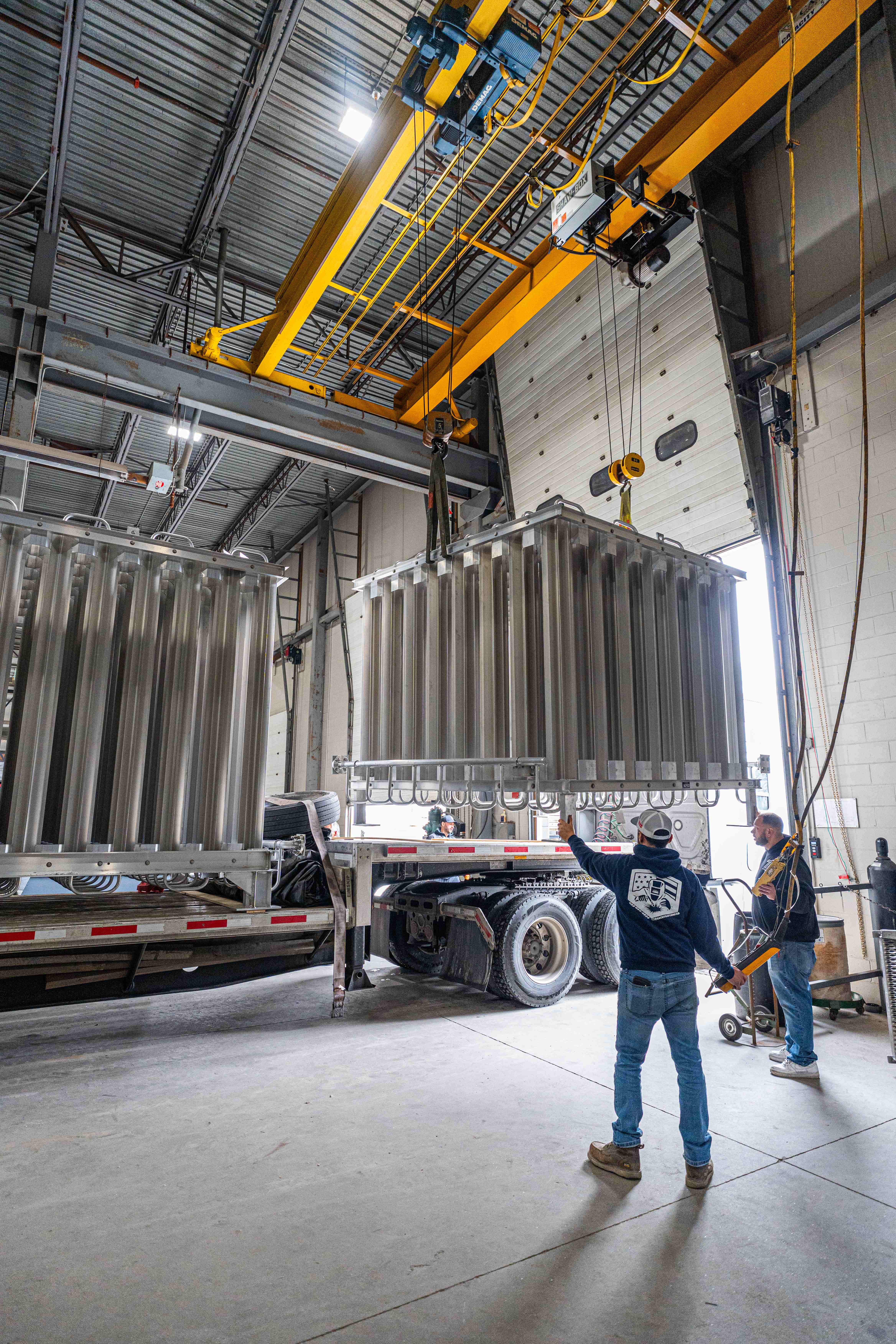 Two FWM workers guiding a finned-aluminum vaporizer bank onto a flatbed under the bridge crane