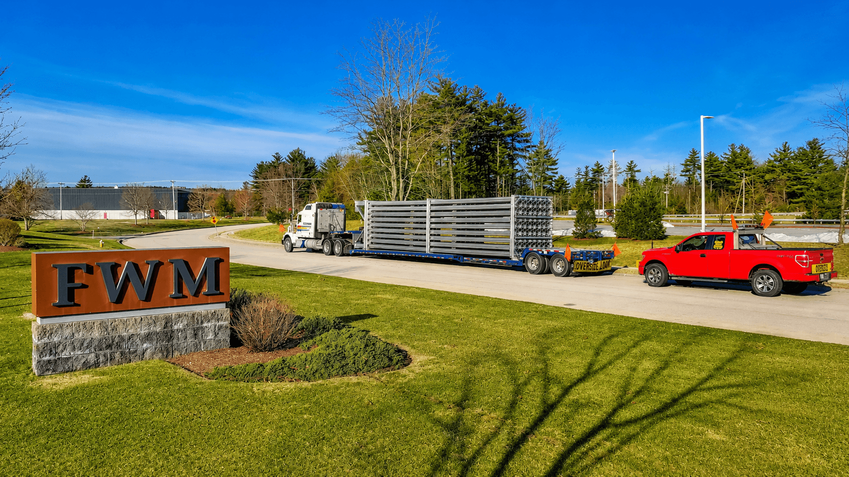 An oversize-load flatbed tractor-trailer hauling racked vaporizer cores past the FWM monument sign at the Hudson, NH campus entrance