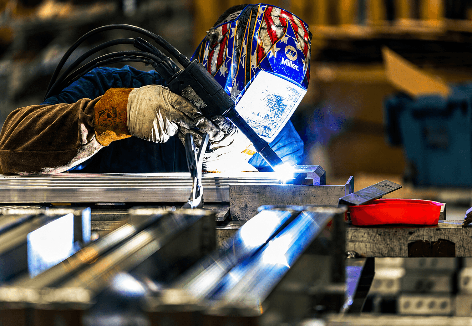 An FWM welder in a stars-and-stripes helmet running a bright arc along aluminum bar stock clamped on a fixture table