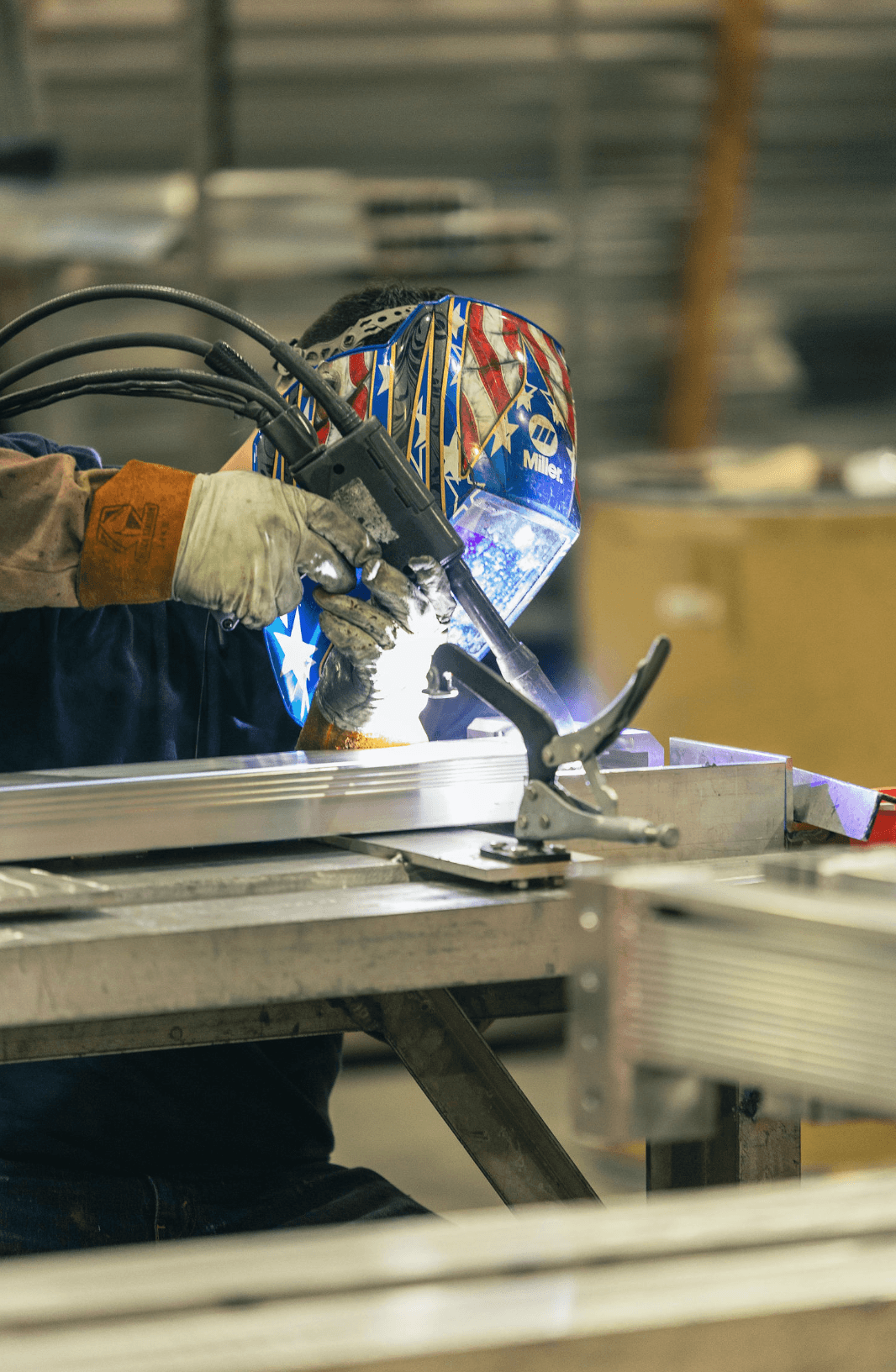 An FWM welder in a Miller welding helmet welding an aluminum extrusion clamped to a fixture on the shop floor