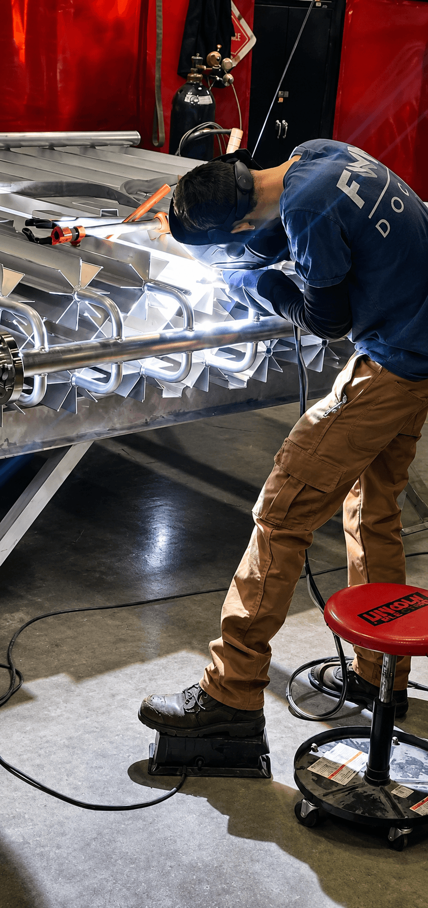 An FWM welder welding a finned-aluminum vaporizer coil assembly on a worktable, weld arc glowing